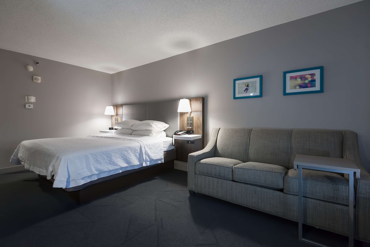 A hotel room with a bed, a sofa, a nightstand with a lamp, and two framed pictures on the wall.