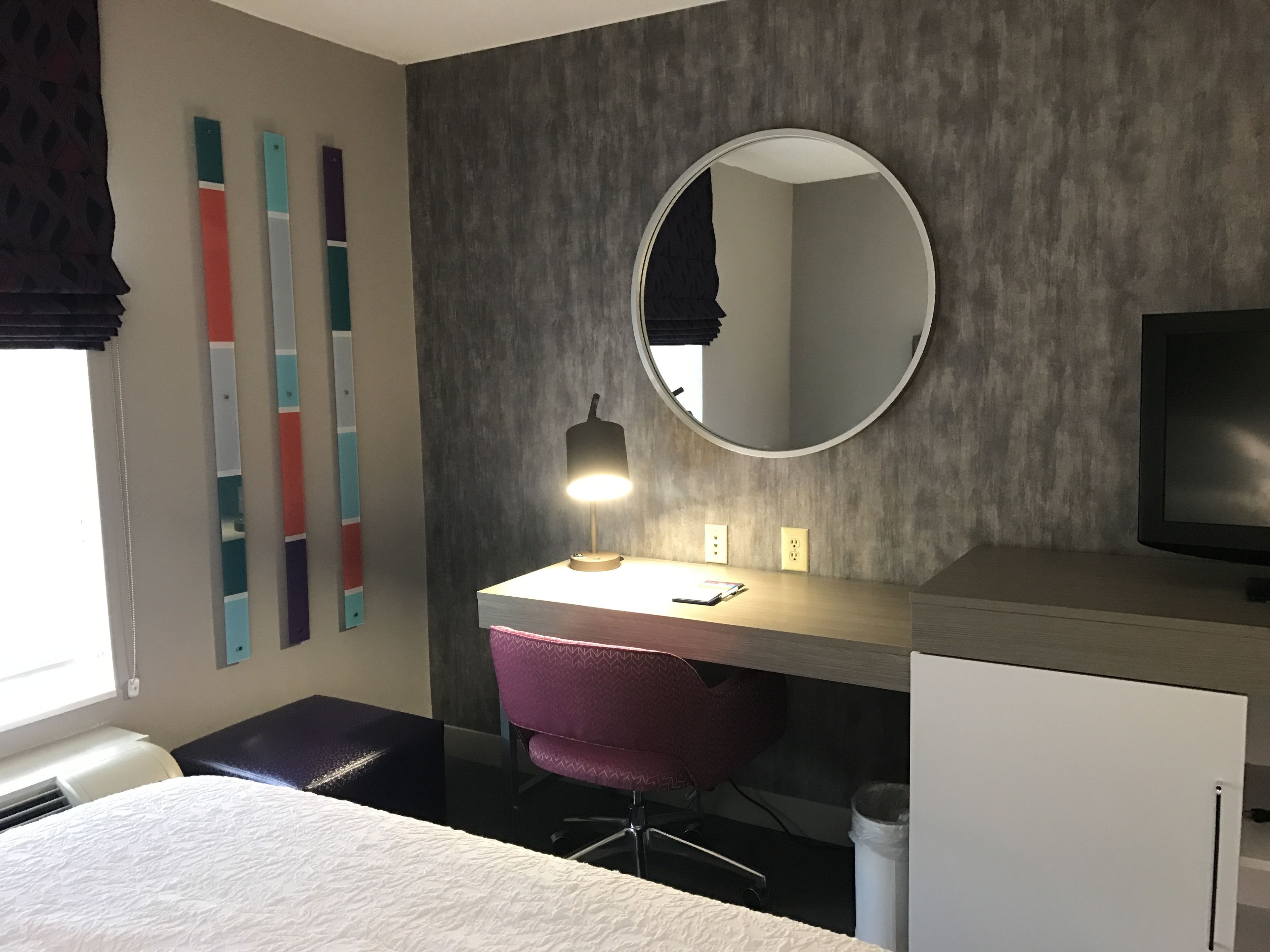 Una habitación de hotel moderna con una cama, un escritorio con lámpara, una silla, un televisor y un espejo.