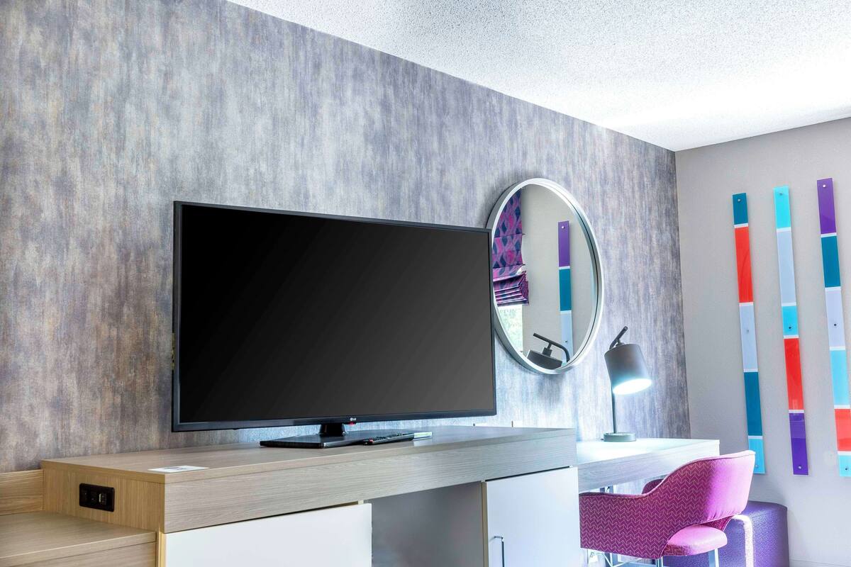 A modern room with a flat-screen TV, a desk, a chair, a round mirror, and colorful wall art.