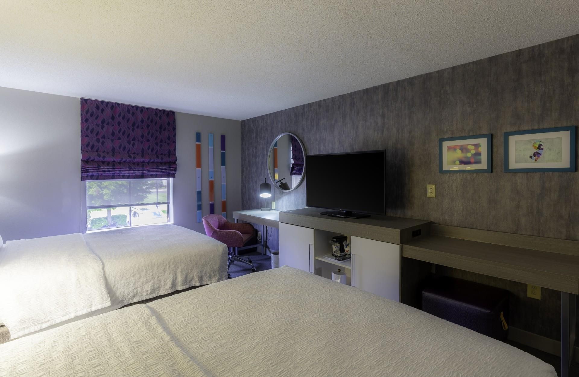 A hotel room with two beds, a desk with a flat-screen TV, a chair, a window with a view, and a wall with a mirror and two framed pictures.