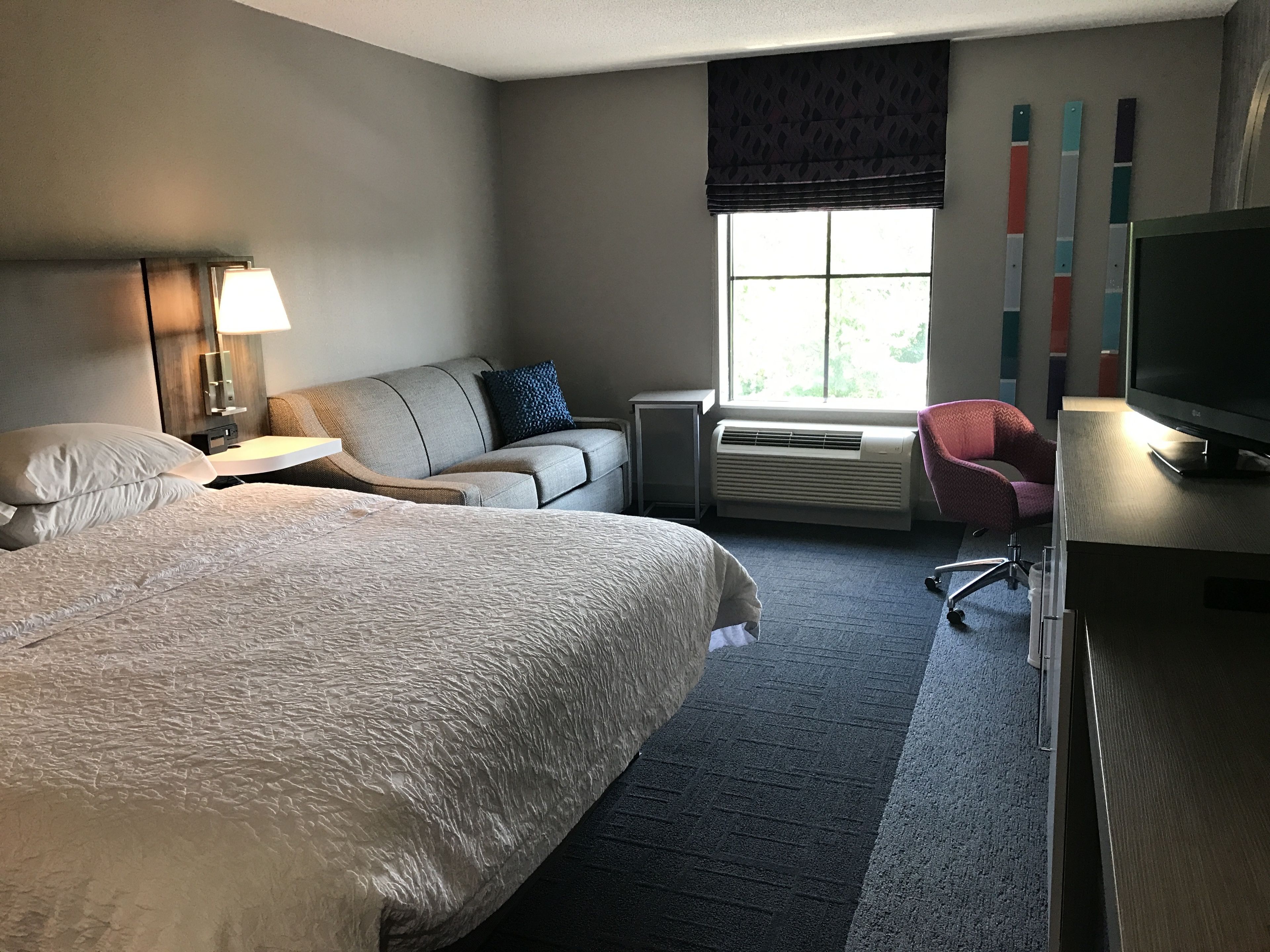 Une chambre d’hôtel avec un grand lit, un canapé, un bureau avec une télévision, une chaise et une fenêtre avec des stores.