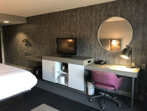 A hotel room with a bed, a desk with a computer, a TV, a mirror, and a lamp.