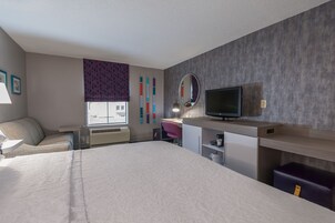 Habitación de hotel con cama, escritorio, silla, televisor y ventana con vistas a edificios.