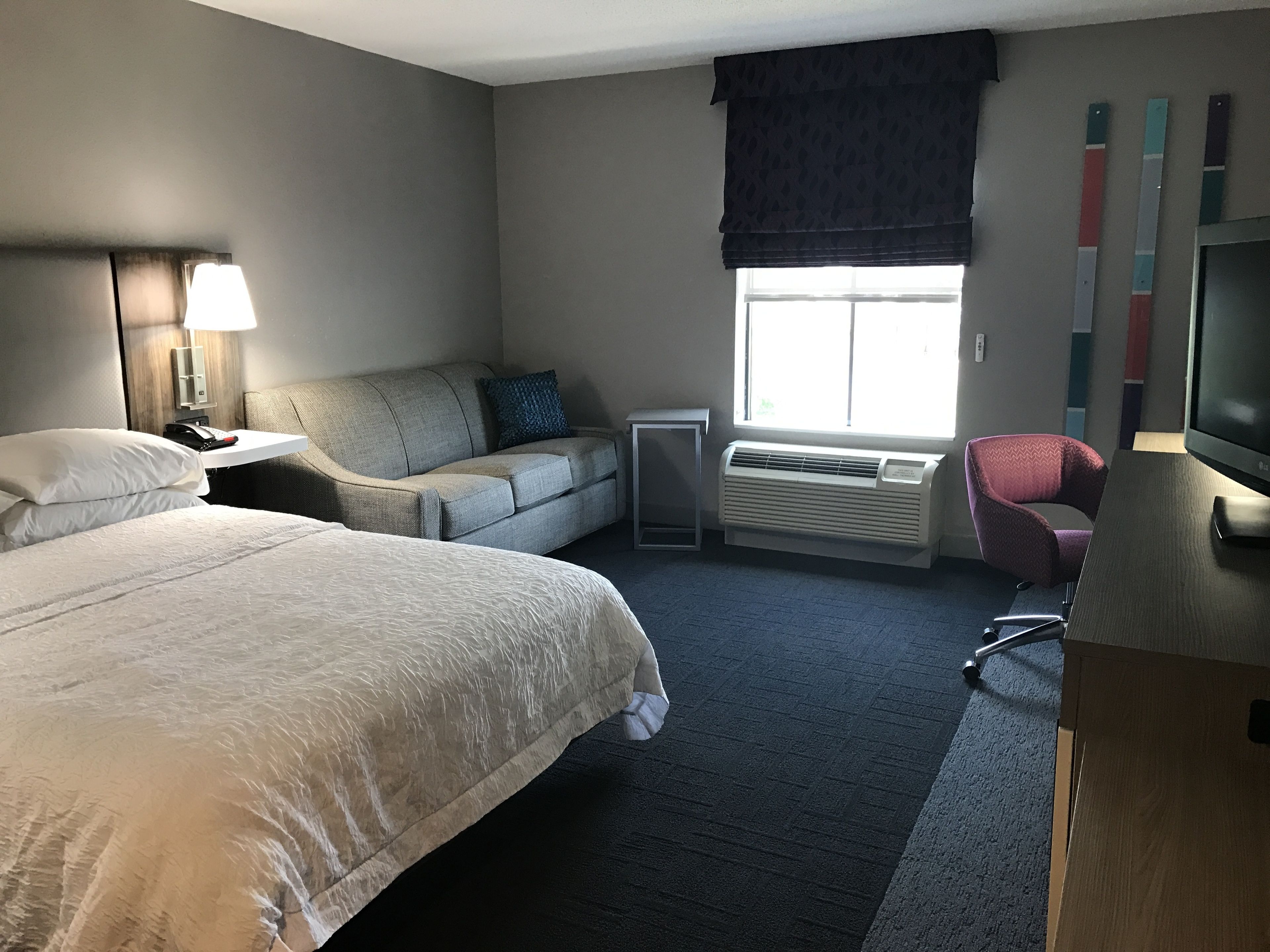 A hotel room with a bed, a sofa, a desk, a chair, a TV, a window with blinds, and a wall-mounted air conditioner.