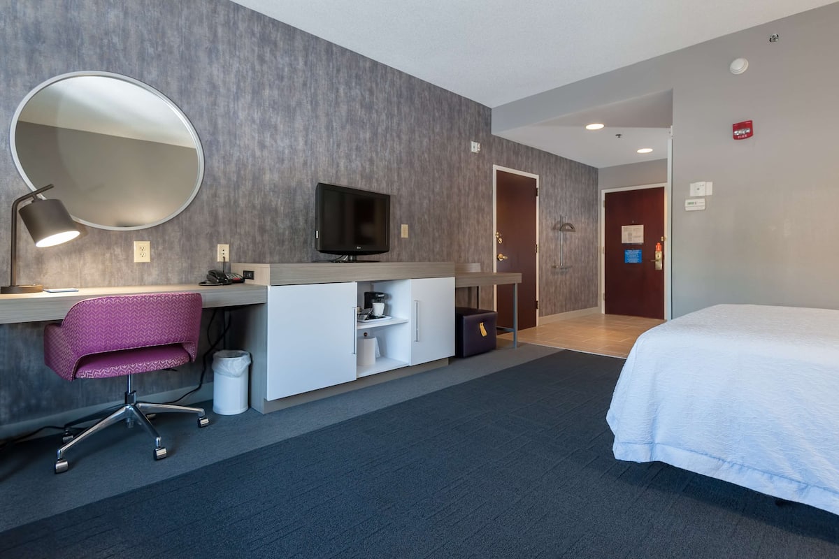 A hotel room with a bed, a desk with a computer, a chair, a TV, and a mirror.
