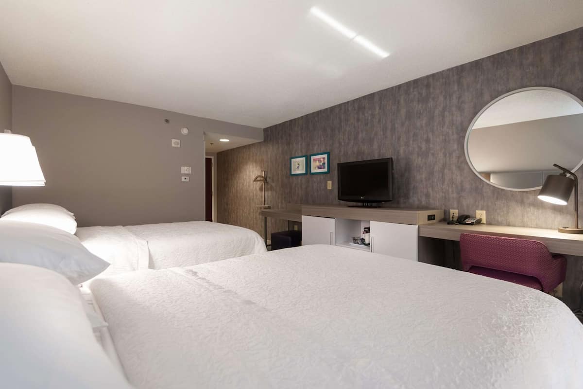 A hotel room with two beds, a desk with a TV, a mirror, and a lamp.