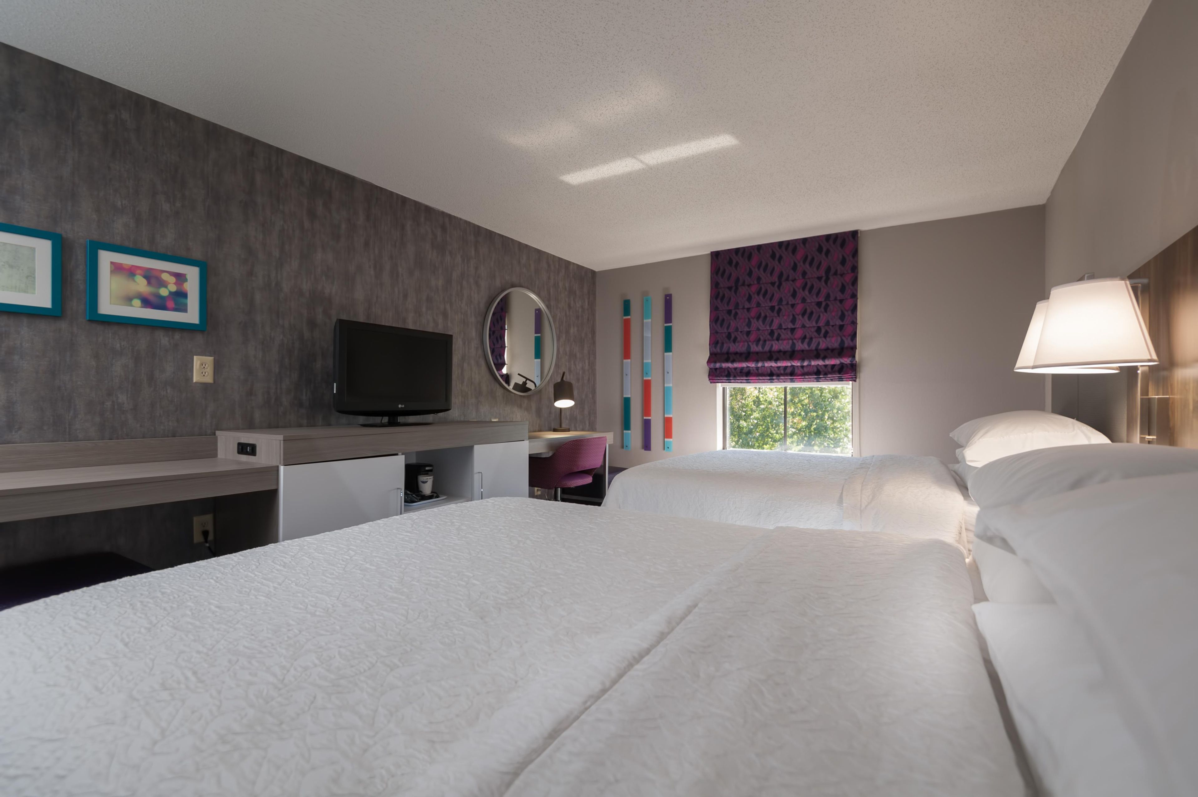 A hotel room with two beds, a desk, a television, a mirror, and a window with curtains.
