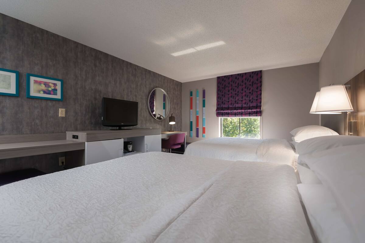 A hotel room with two beds, a desk with a computer, a TV, a mirror, and a window with a view of trees.