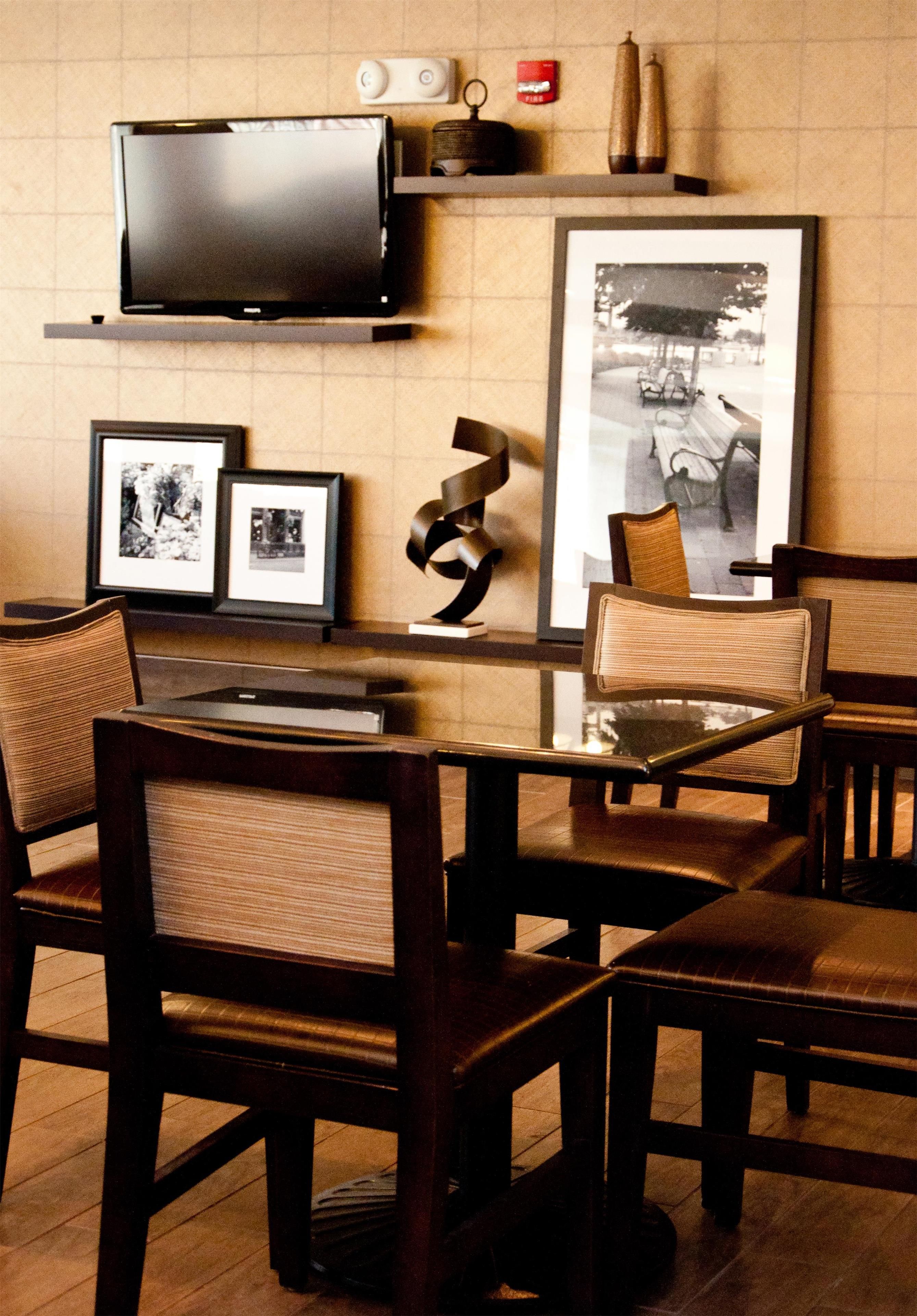 A dining area with a glass table, wooden chairs, a television mounted on a shelf, and framed pictures on the wall.