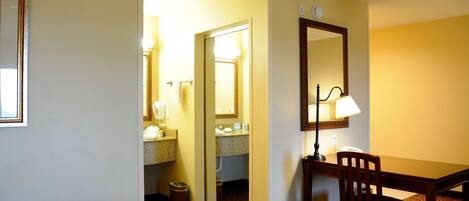 Habitación de hotel con cama, escritorio con silla, espejo y baño visible a través de una puerta abierta.