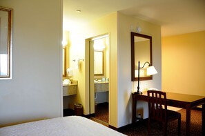 A hotel room with a bed, a desk with a chair, a mirror, and a bathroom visible through an open door.