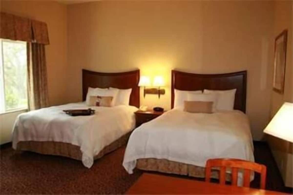 Habitación de hotel con dos camas, una mesita de noche con lámpara y una ventana con cortinas.