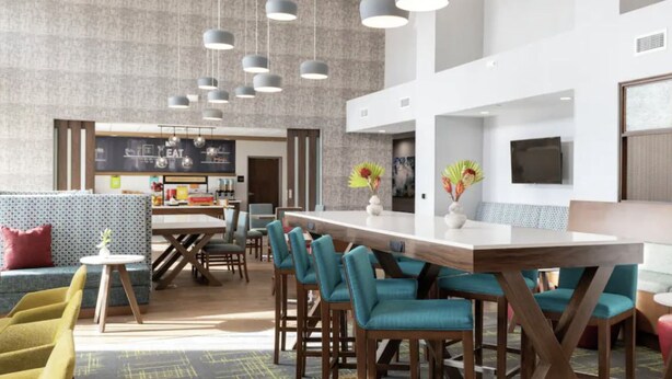 A modern hotel lobby with a dining area, seating arrangements, and a kitchen in the background.
