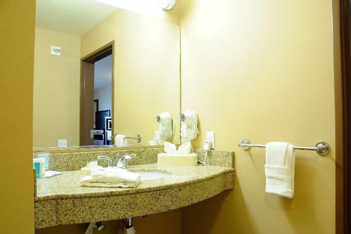 Baño de hotel con lavamanos de encimera de granito, un espejo, toallero y una puerta visible que da a otra habitación.