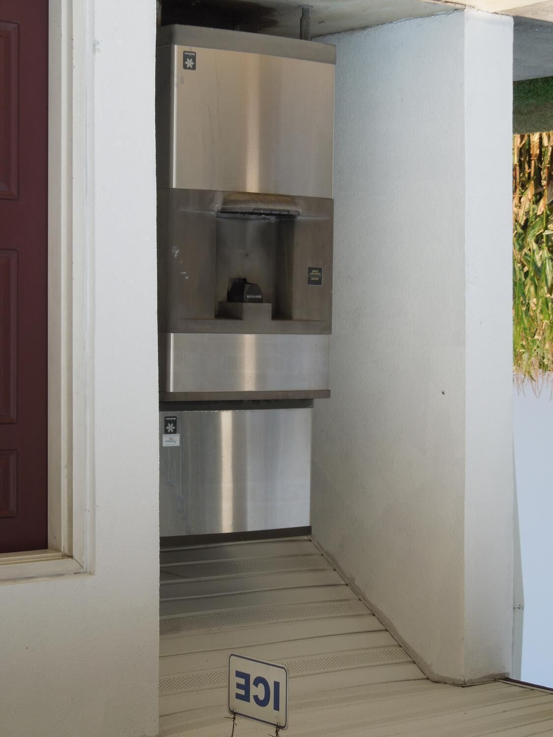 A compact kitchen with stainless steel appliances, a red door, and a small window with a view of corn plants.