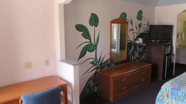 A hotel room with a wooden desk, a chair, a dresser, a television, and a large plant. - Travelers Inn and Suites Liberty (Liberty)