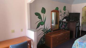 A hotel room with a wooden desk, a chair, a dresser, a television, and a large plant.