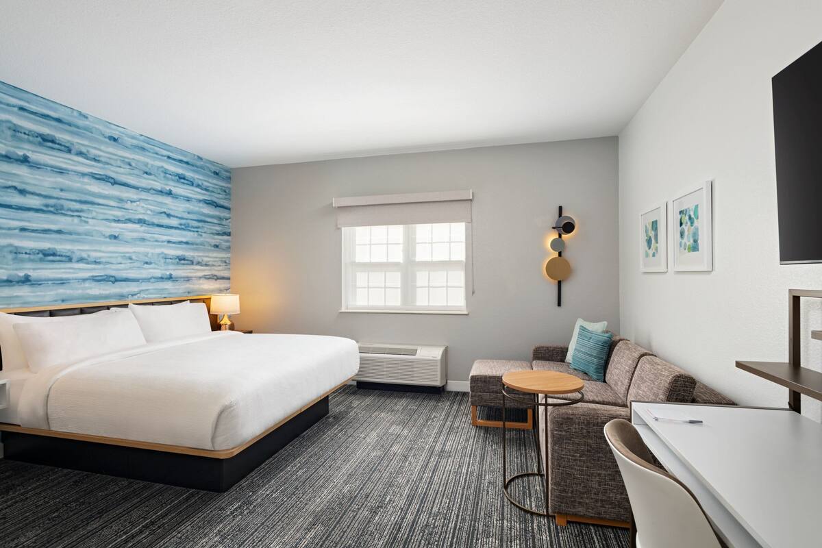 Ein Hotelzimmer mit einem großen Bett, einer Couch, einem kleinen Tisch und einem Schreibtisch. Es gibt ein Fenster mit Blick, eine Wanduhr und ein Wandbild.