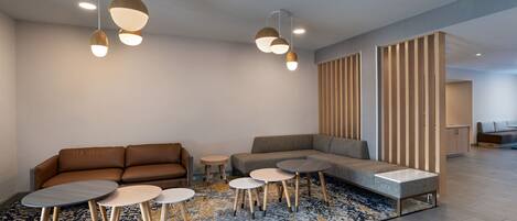 Una sala moderna con un sofá marrón, un sofá modular gris y un tapete estampado. Hay cuatro mesas redondas con patas de madera.