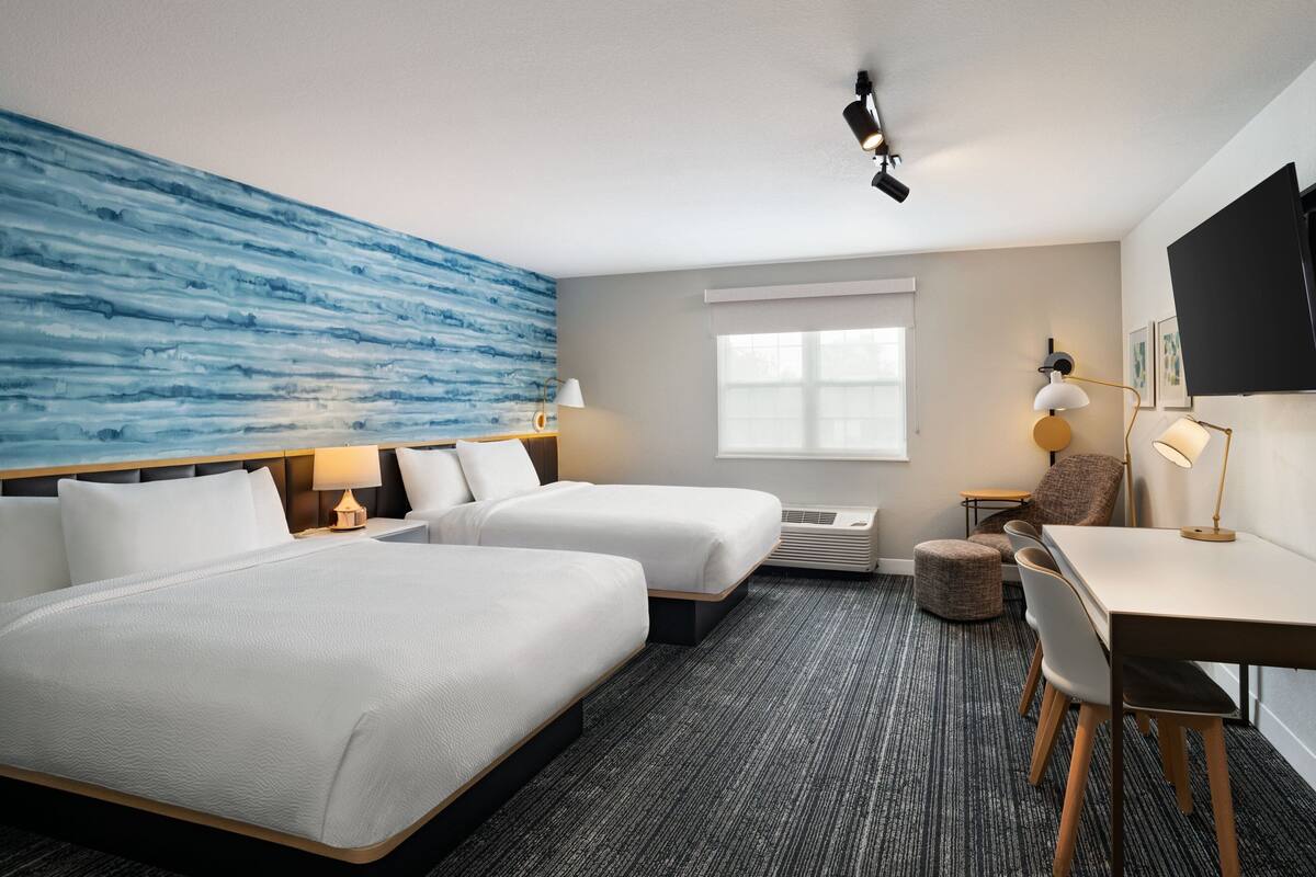 Ein Hotelzimmer mit zwei Betten, einem Schreibtisch, einem Sessel, einem Fernseher und einem großen Fenster.