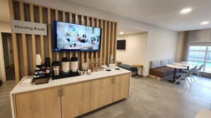 Un área de desayuno moderna con una barra de madera, un televisor mostrando un programa matutino y un espacio de estar con mesas y sillas.