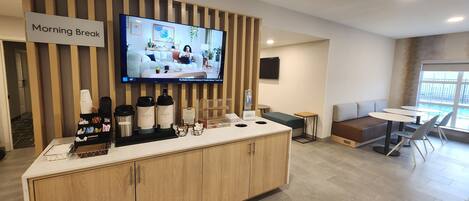 A modern breakfast area with a wooden counter, a television displaying a morning show, and a seating area with tables and chairs.