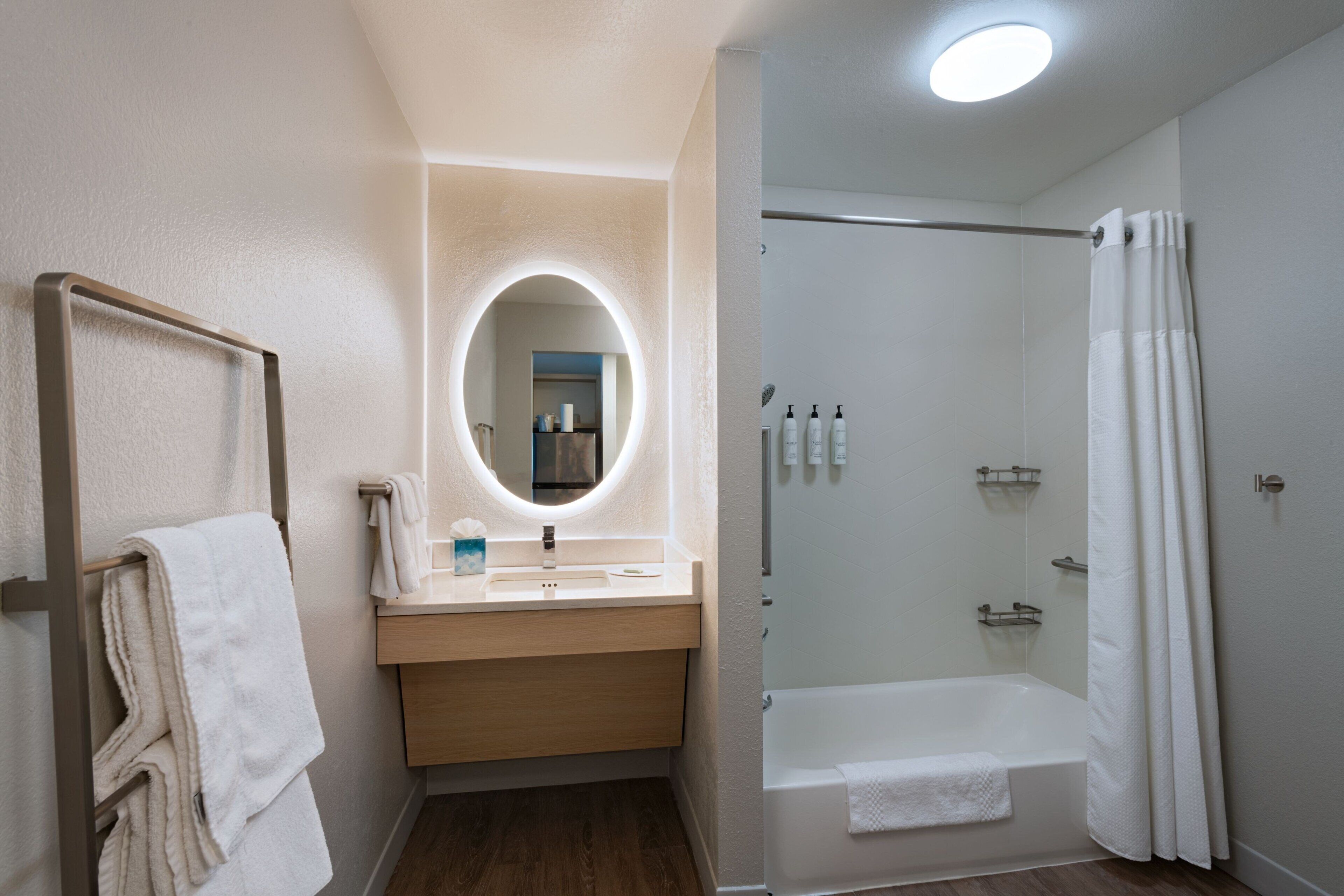 A modern bathroom with a wooden vanity, a round mirror, a bathtub, and a glass-enclosed shower.