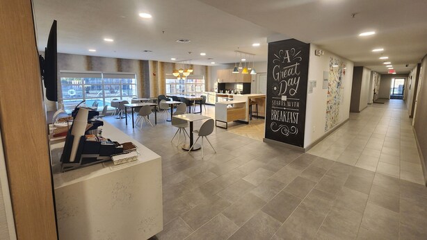 Un comedor moderno con varias mesas y sillas, una zona de cocina y un largo pasillo con un mural motivacional.