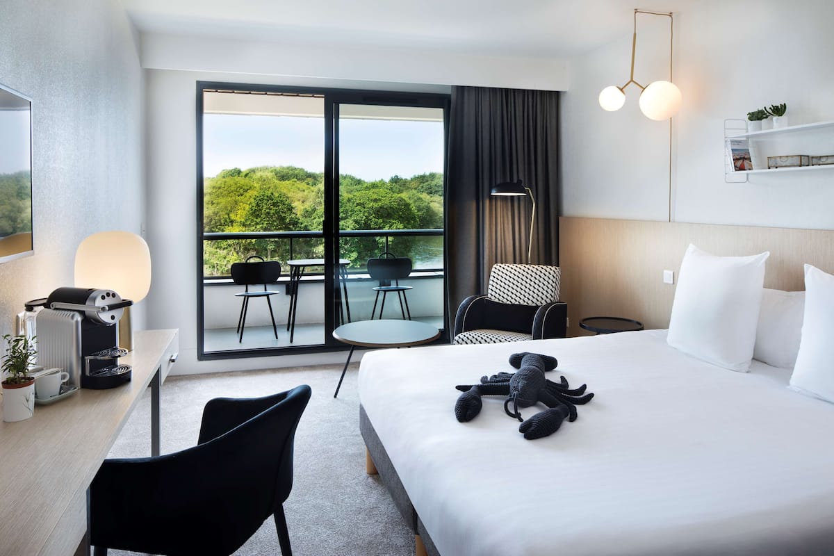 A hotel room with a large bed, a desk with a coffee maker, a chair, a small table, and a balcony with a view of greenery.
