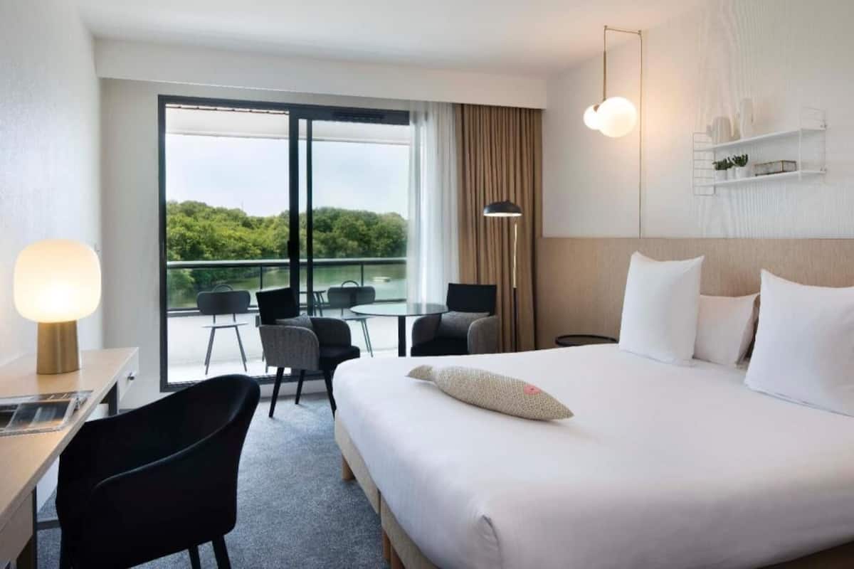 A hotel room with a bed, a desk, a chair, a table, a lamp, a balcony with chairs, and a view of greenery.
