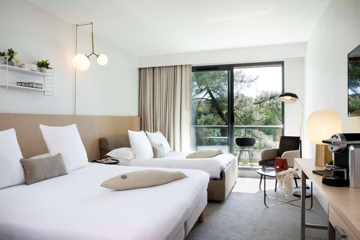 A hotel room with two beds, a desk with a coffee machine, a chair, and a large window with a view of trees.