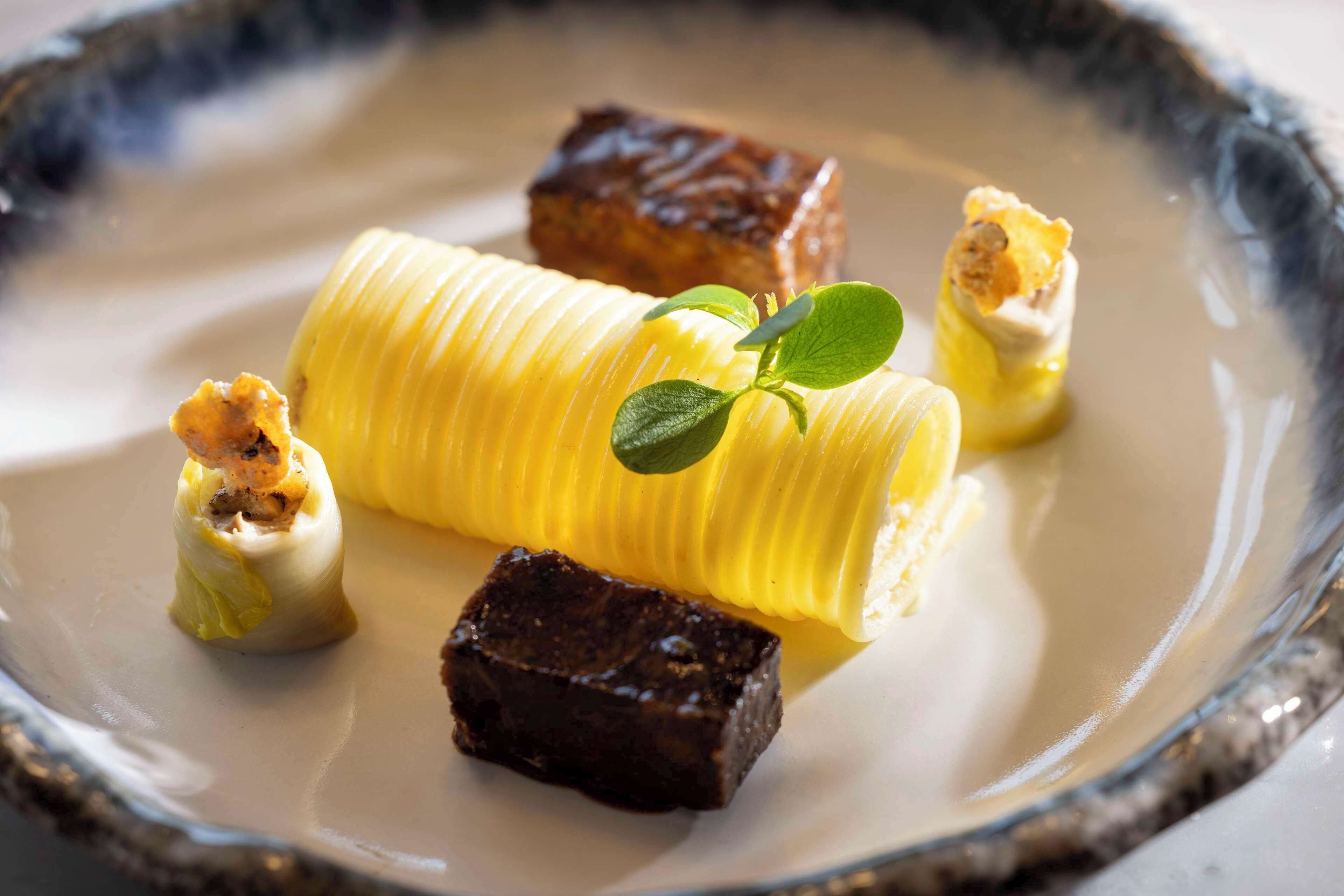 Un plato de postre con una base cremosa, un objeto amarillo cilíndrico y varios trozos marrón oscuro.