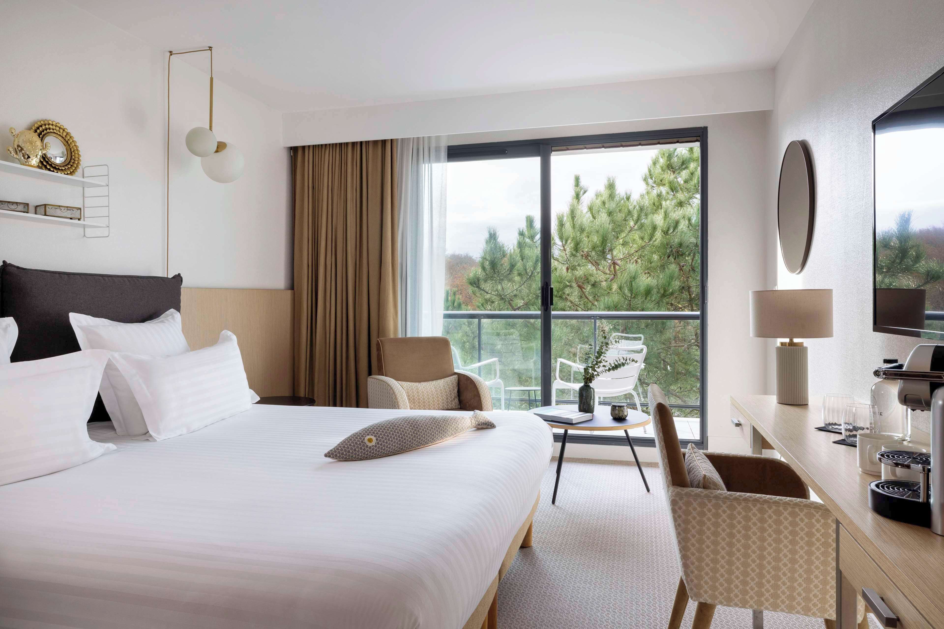 A hotel room with a large bed, a desk with a chair, a television, and a window with a view of trees.