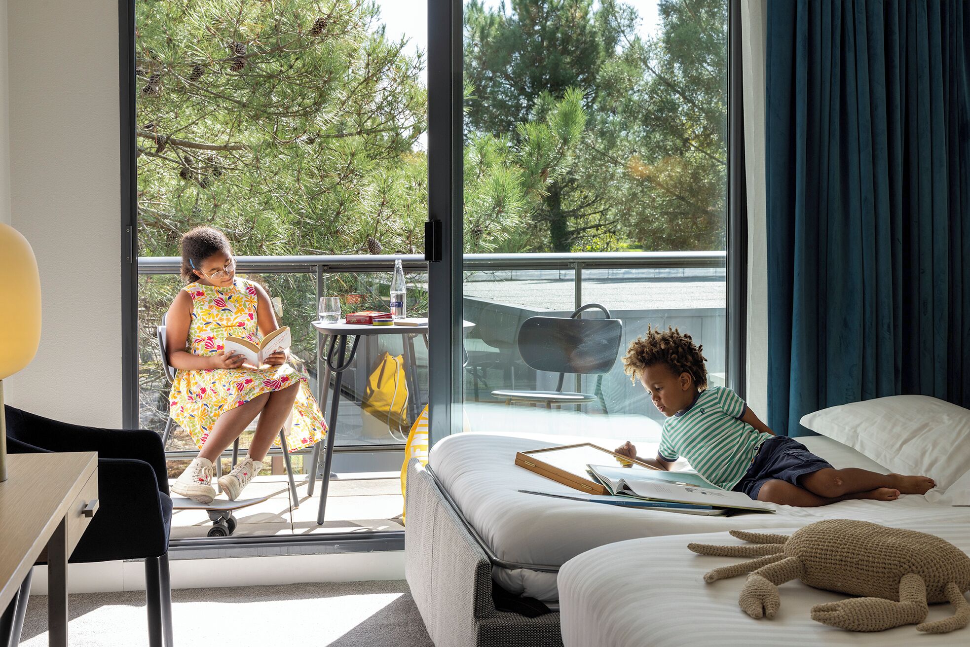 A room with a bed, a window with a view of trees, a table with a chair, and a person reading a book.