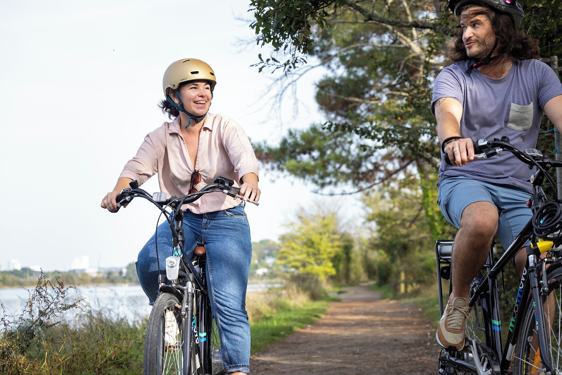Due persone in bicicletta su un sentiero, sullo sfondo alberi e uno specchio d'acqua.
