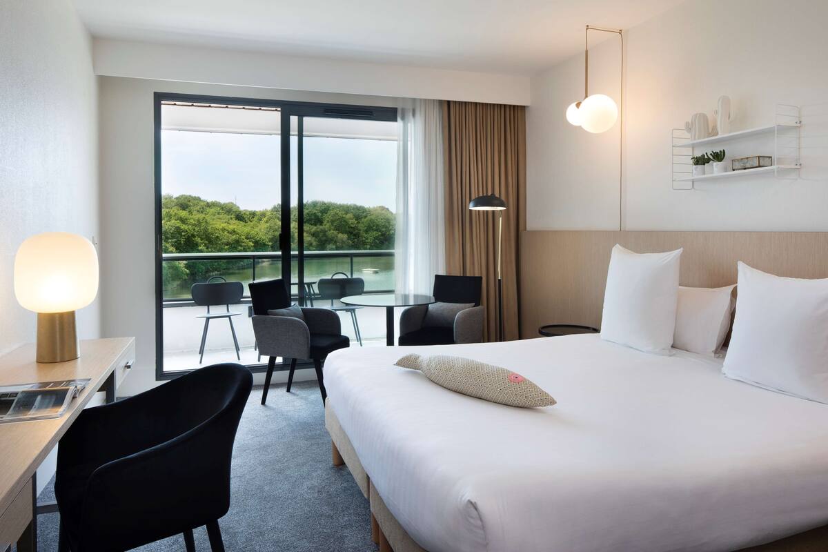 A hotel room with a large bed, a desk, a chair, a table, and a view of greenery through the window.