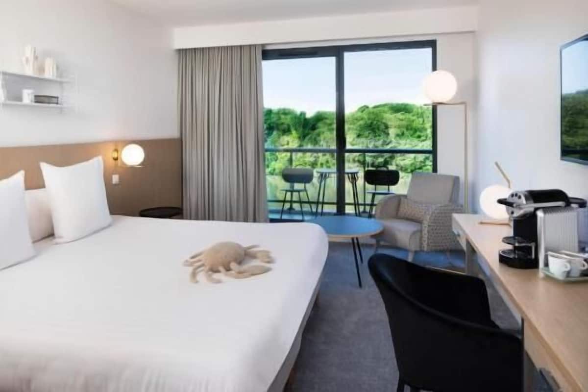 A hotel room with a bed, a desk, a chair, a small table, a lamp, and a view of greenery through the window.