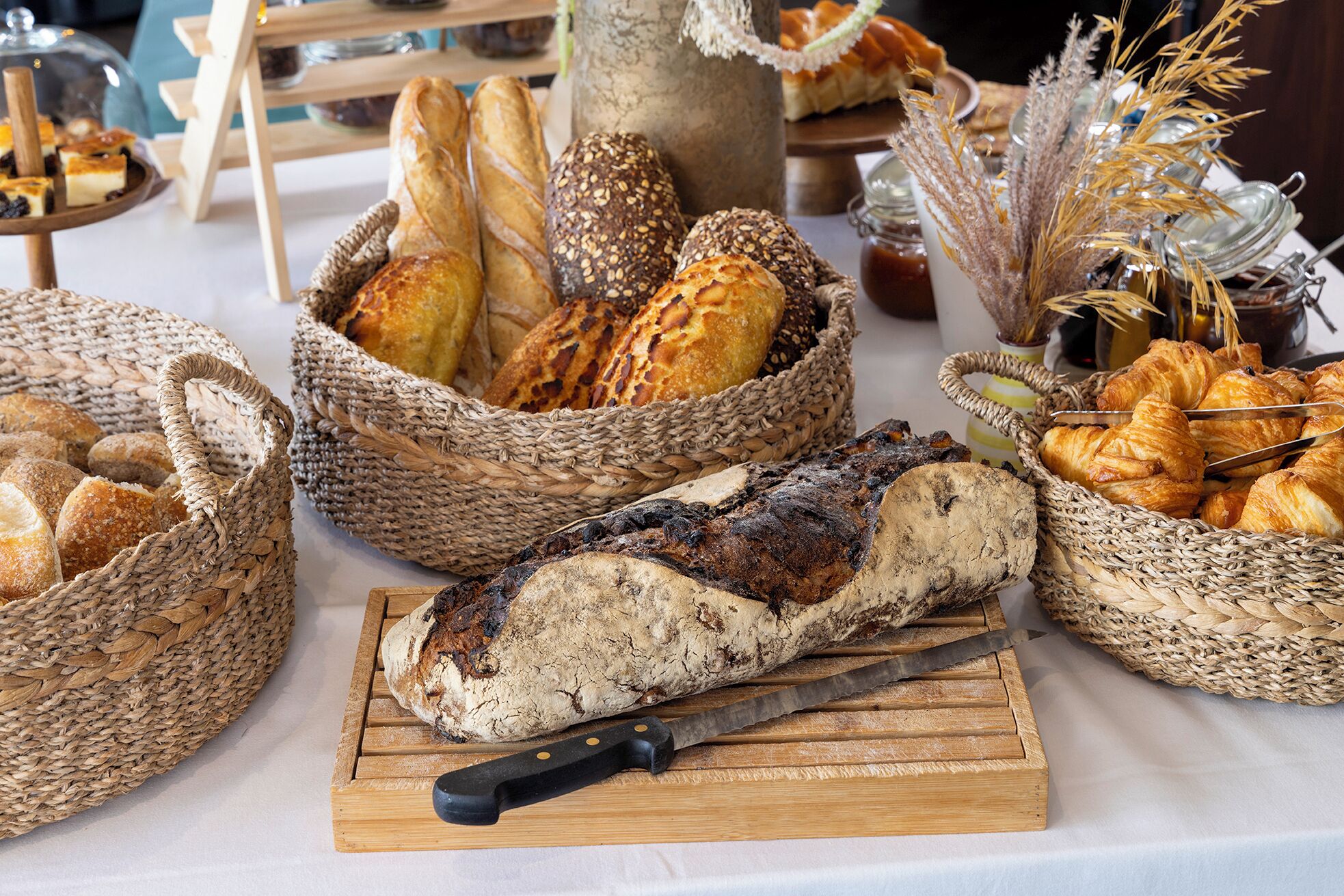 Variedad de panes y pasteles expuestos sobre una mesa con cestas y tabla de cortar.