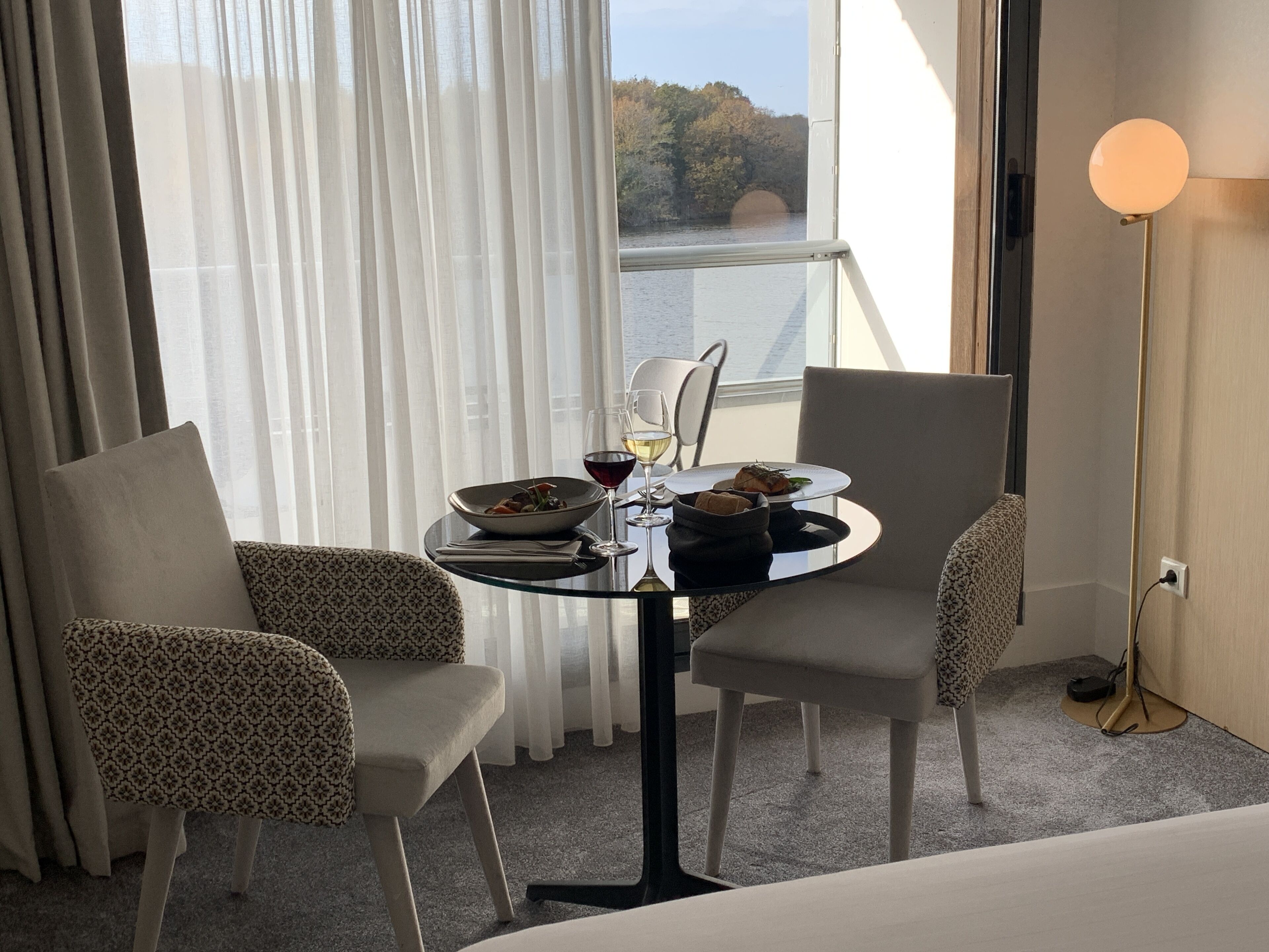A hotel room with a dining table set for two, a view of a bridge and trees, and a lamp on the side.