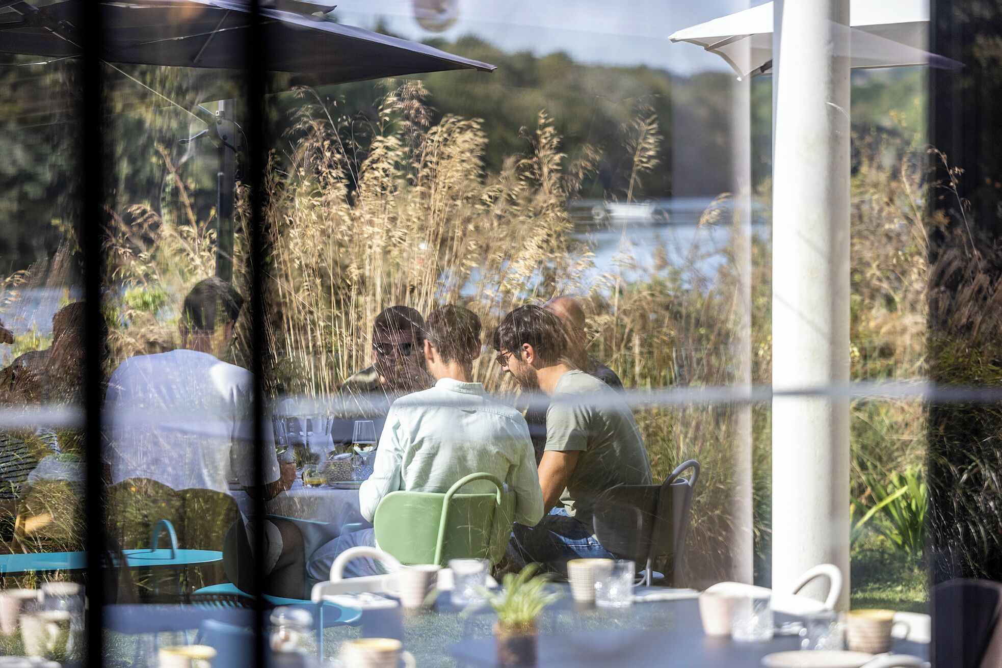 A dining area with tables and chairs, a view of tall grasses and trees, and a body of water in the background.