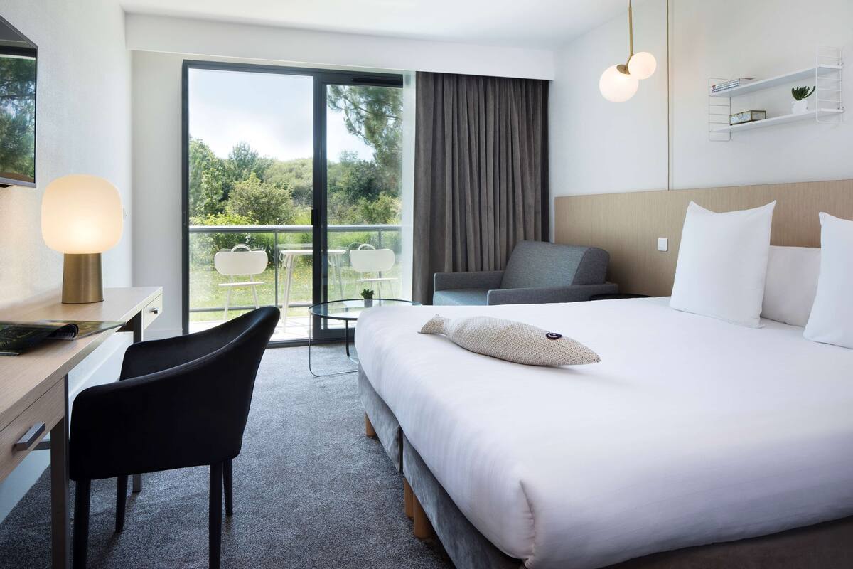 A hotel room with a large bed, a desk with a chair, a balcony with a table and chairs, and a view of greenery outside.