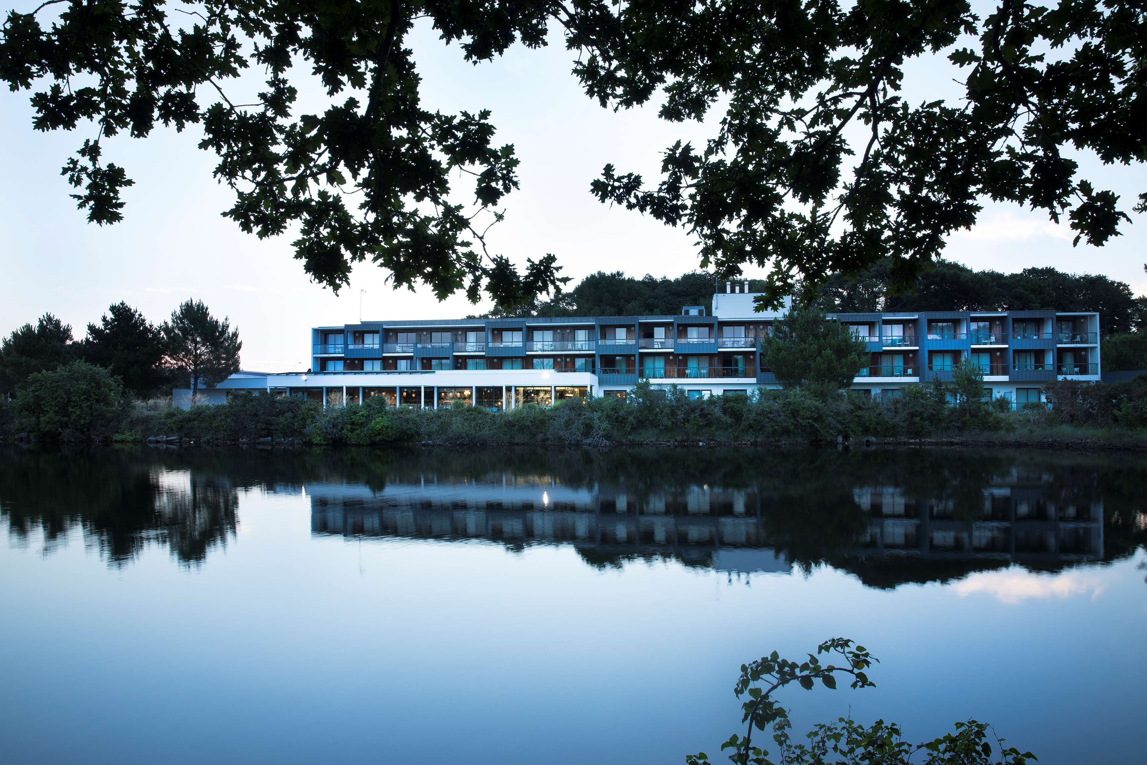 A multi-story hotel with a modern design, located near a body of water, featuring balconies and large windows.