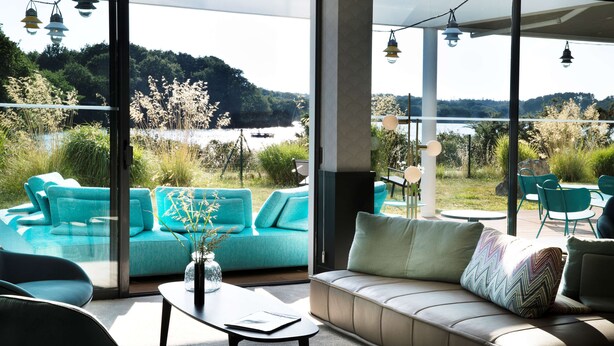 A modern outdoor seating area with a sofa and chairs, a table with a vase, and a view of a lake and hills. - Best Western Plus Hotel Les Rives Du Ter (Larmor-Plage)