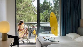 A hotel room with a bed, a desk with a lamp, a chair, a table with a surfboard, and a view of trees and sky through the window.