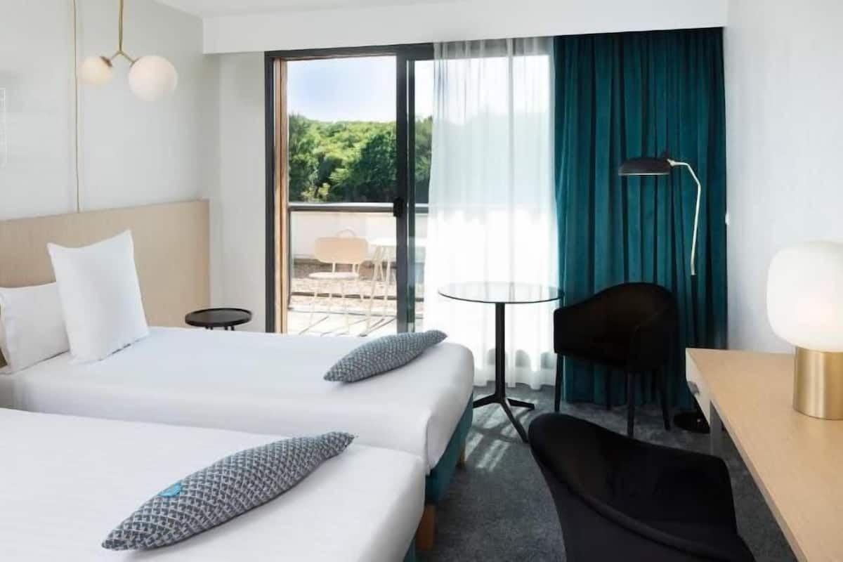 A hotel room with two beds, a desk, a chair, a table, a lamp, and a view of the outdoors.