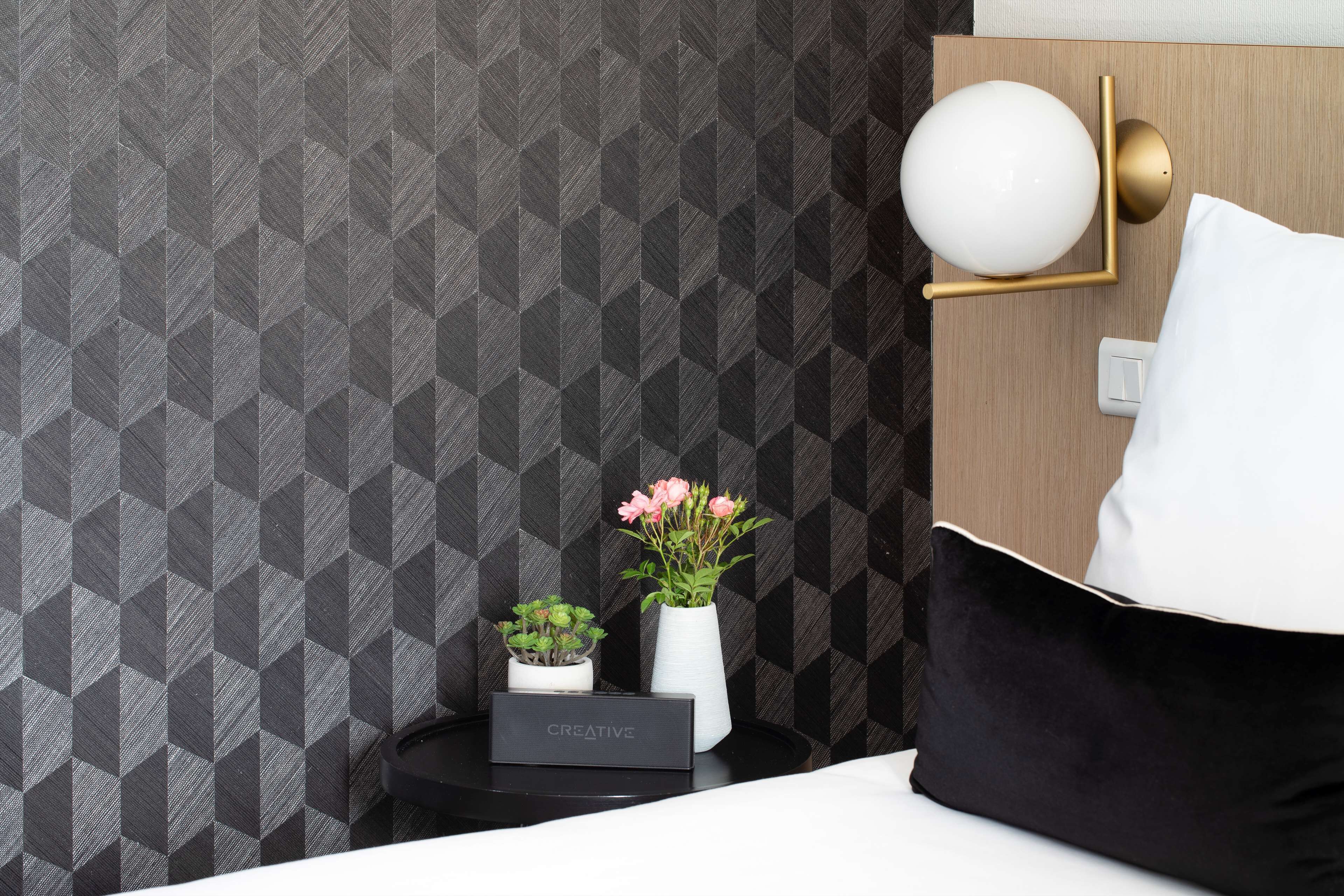 A neatly arranged hotel room with a geometric-patterned wallpaper, a bedside lamp, a small table with a box labeled "CREATIVE," and two potted plants.