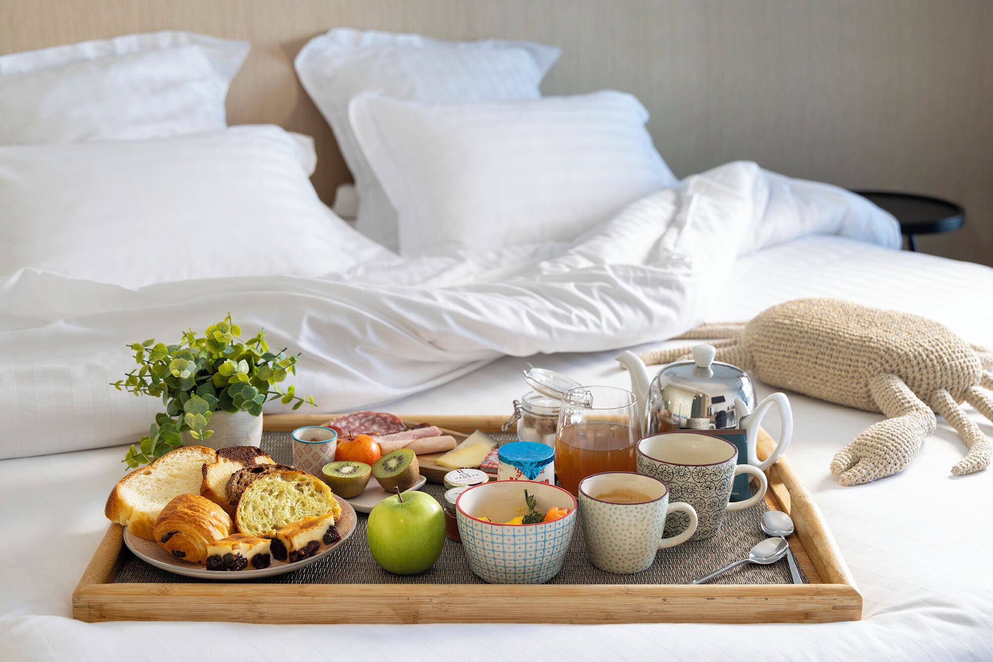 A breakfast tray with toast, croissants, kiwi, an apple, and a bowl of cereal on a bed with white linens and pillows.