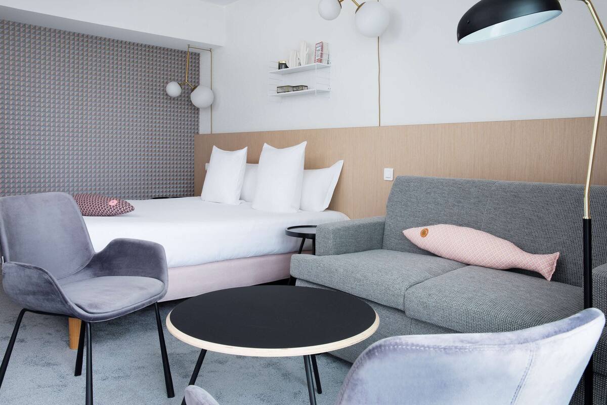 A modern hotel room with a bed, grey armchairs, a round black table, and a black floor lamp.