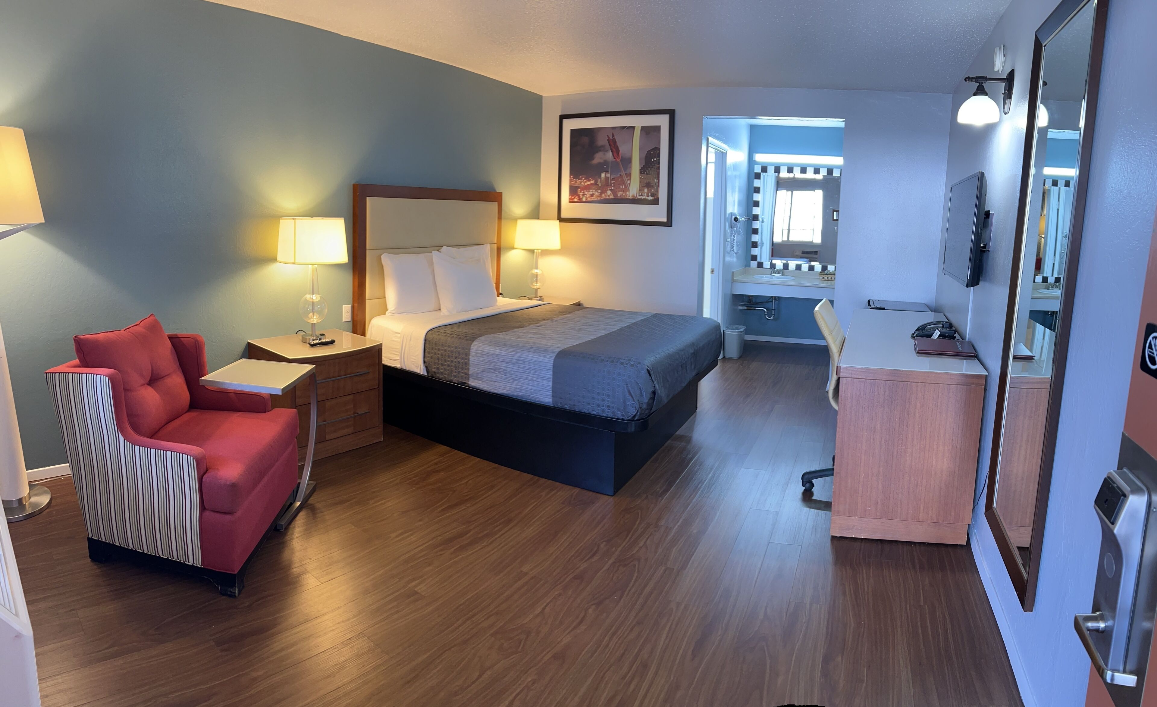 A hotel room with a bed, a desk, a chair, a mirror, and a bathroom visible through an open door.