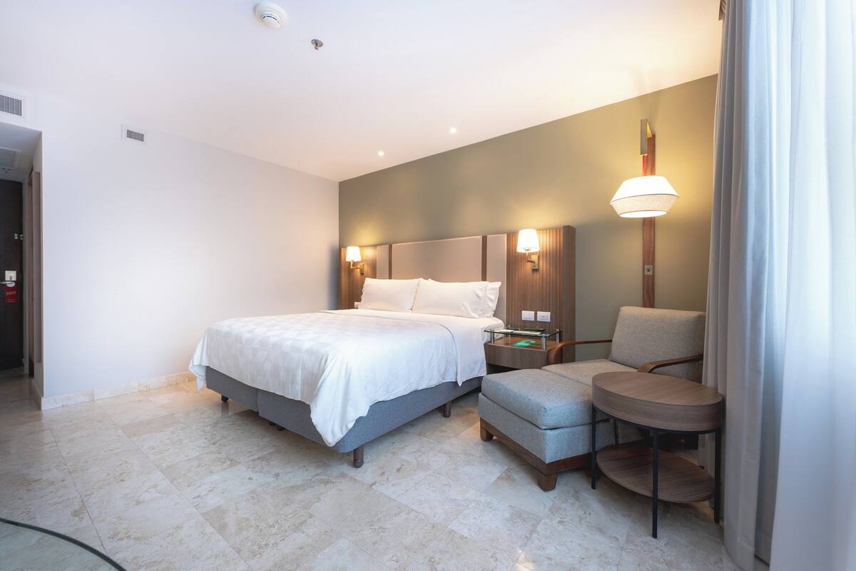 A hotel room with a bed, a chair, a small table, and a lamp.